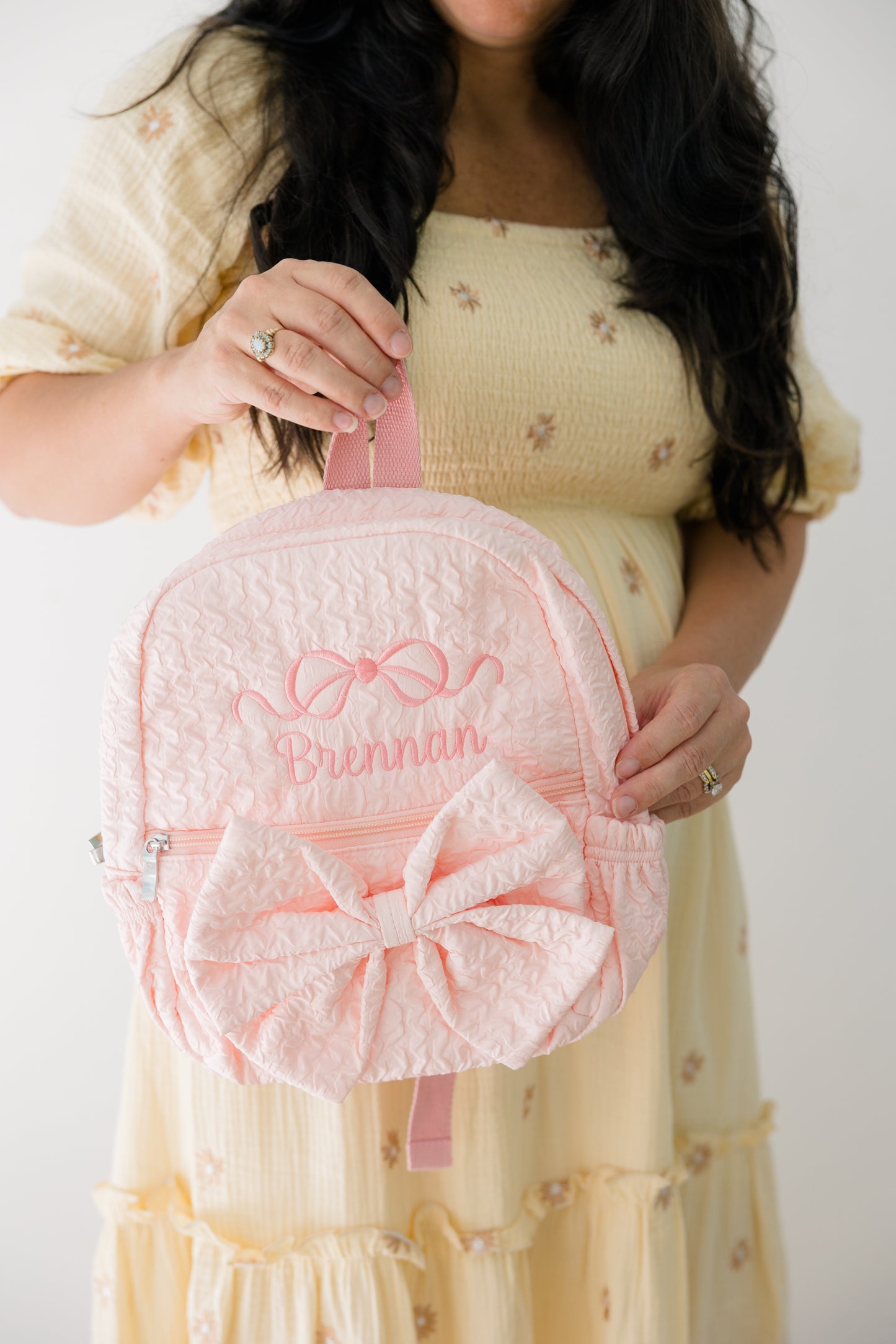 Person holding a pink backpack with a large bow and embroidered name against a plain background