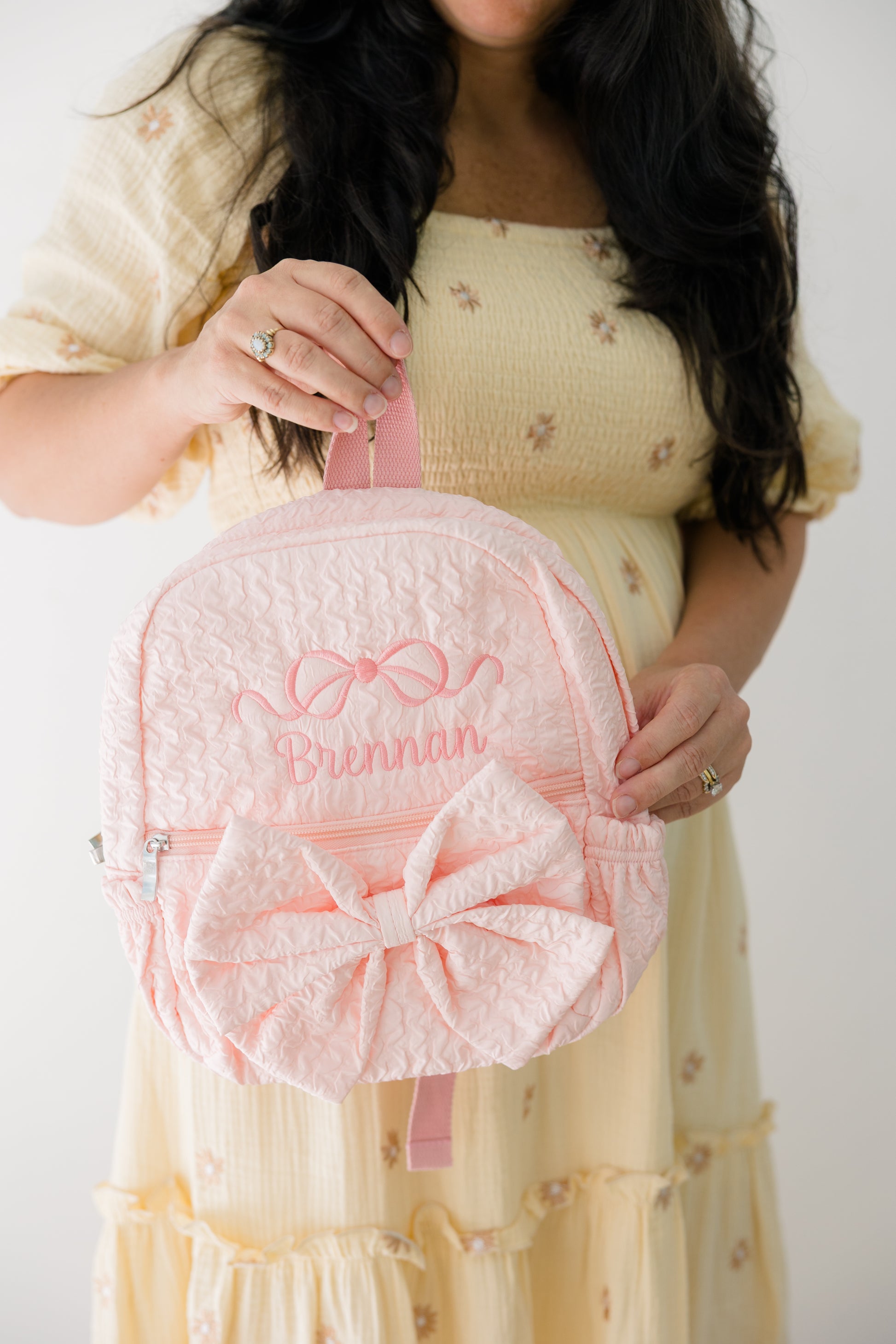Person holding a pink backpack with a large bow and embroidered name against a plain background