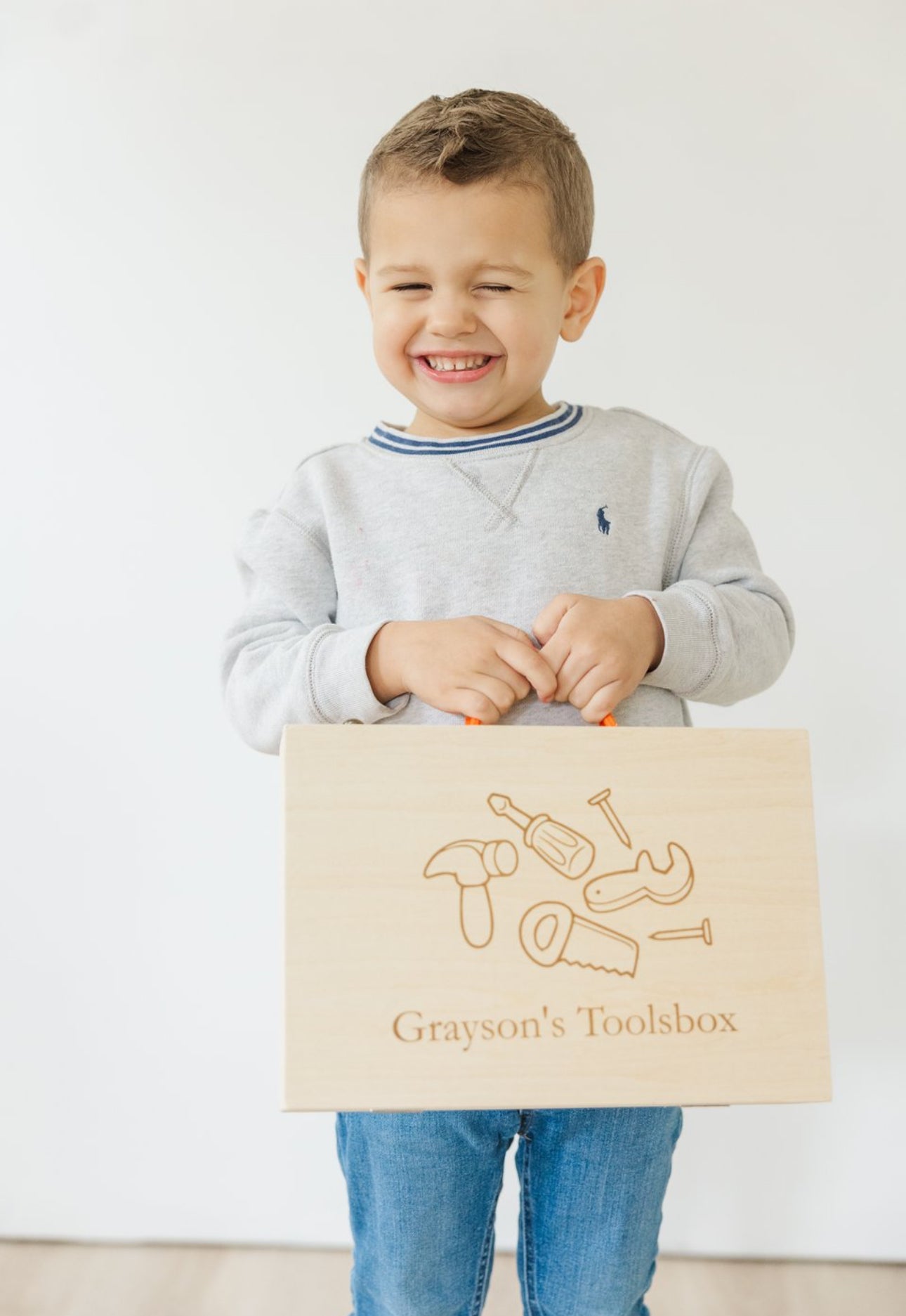 A young child standing, holding a personalized wooden toolbox with a handle. The toolbox has an engraved name 'Aaron' on the front and features a design with various tools.