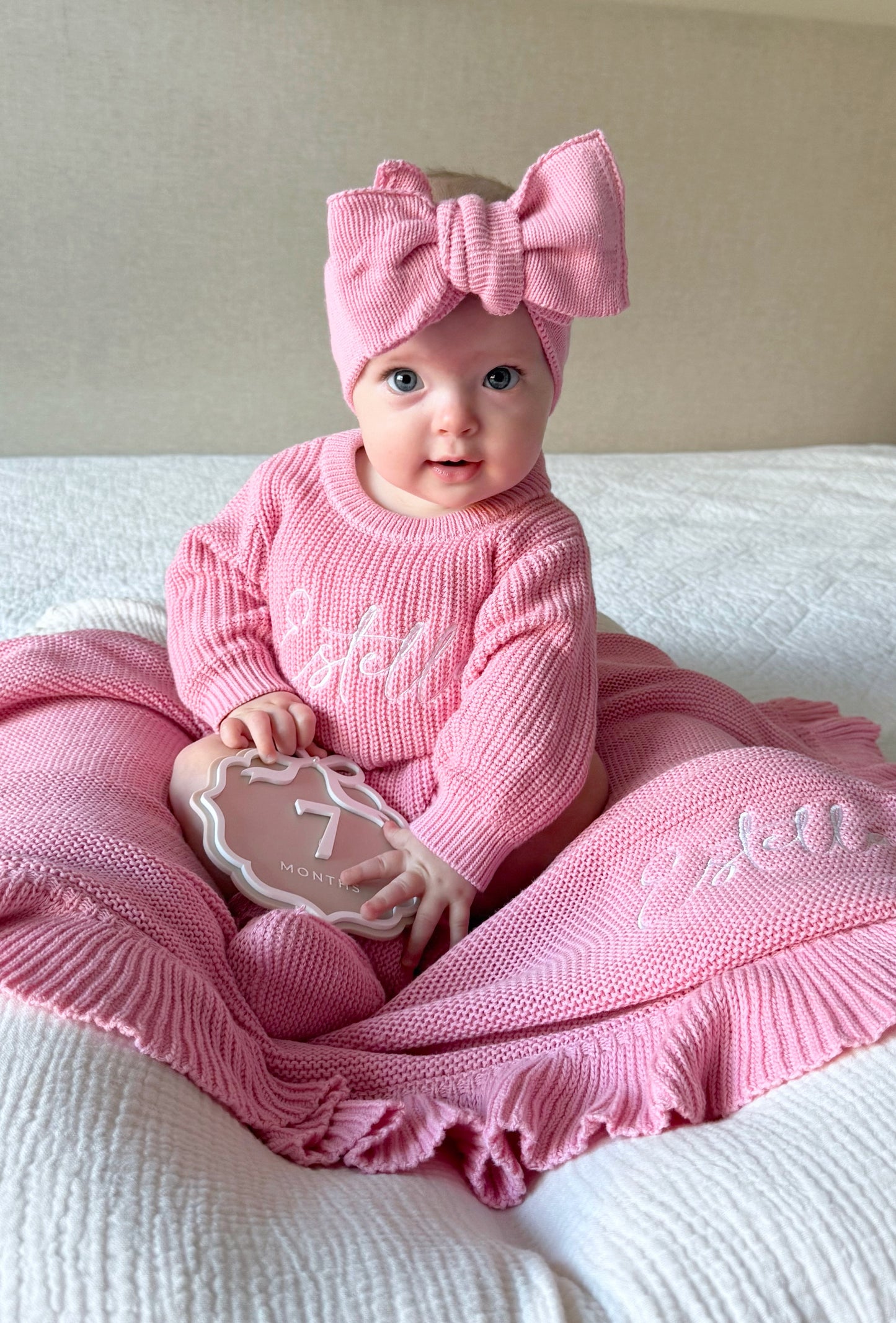 Baby in pink outfit with a large bow, sitting on a white blanket.