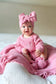 Baby in pink outfit with a large bow sitting on a pink blanket.