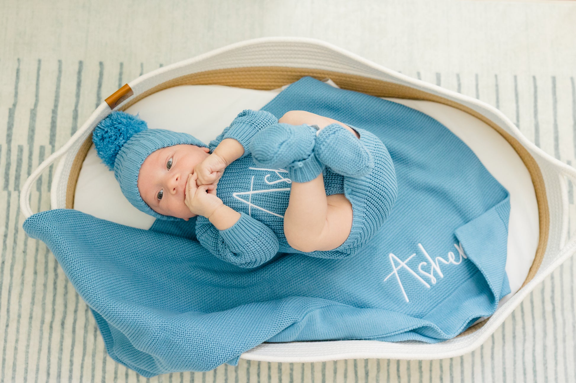 Newborn baby in blue outfit and hat lying on a blue blanket with 'Asher' embroidered, in a white basket.