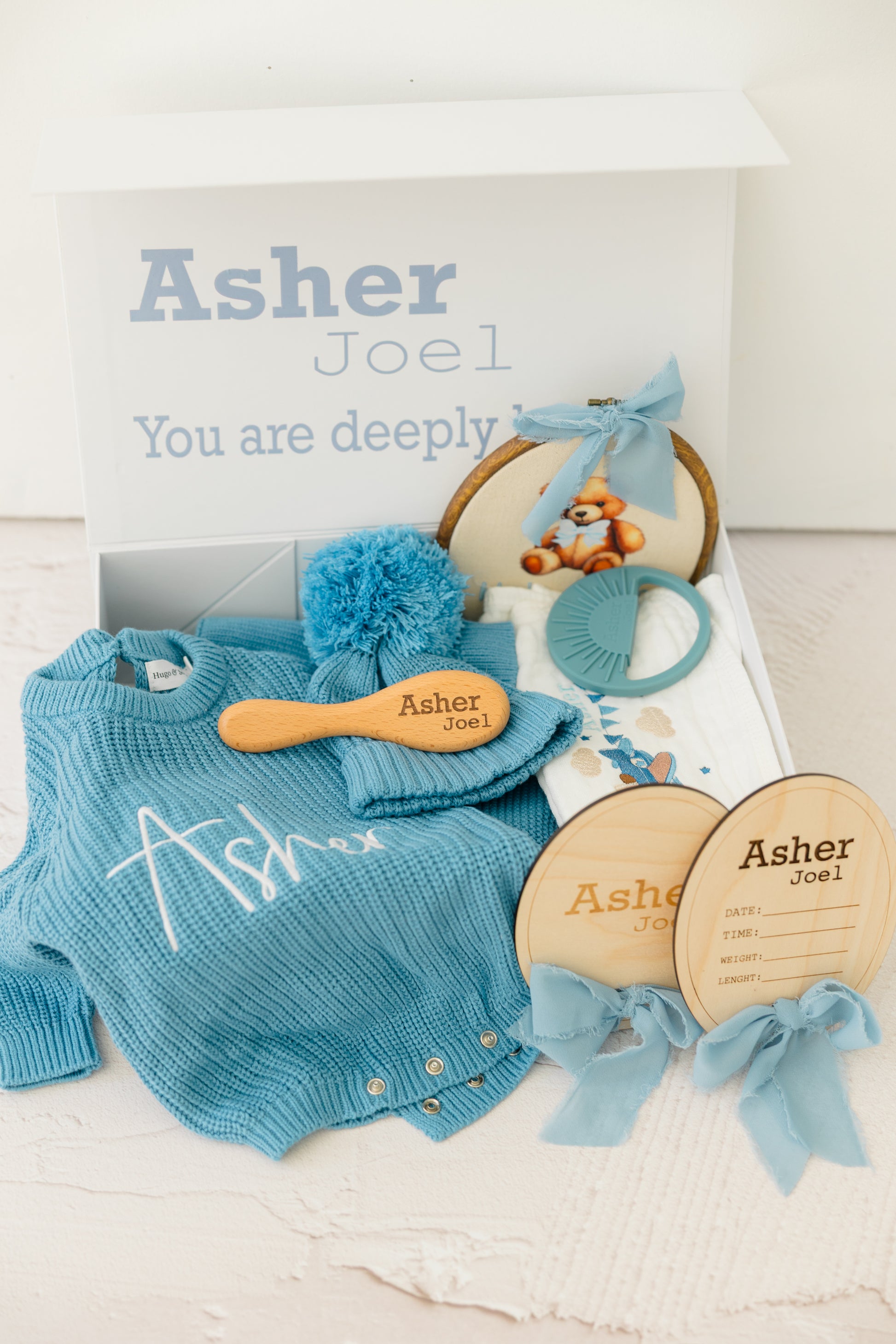 Baby shower gift set with blue baby outfit, wooden toys, and personalized cards on a white background.
