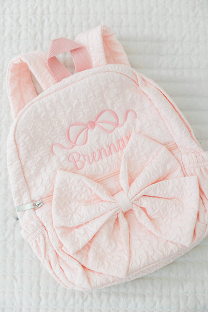 Pink backpack with bow and embroidered name on a textured surface