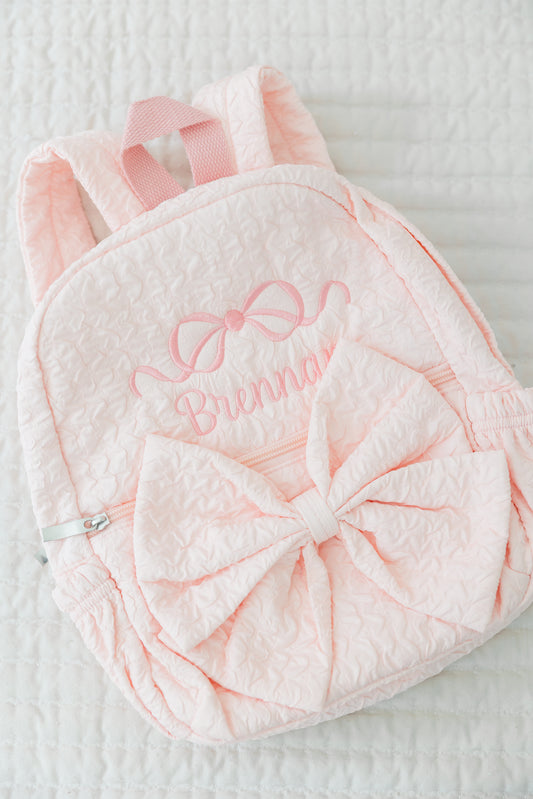 Pink backpack with bow and embroidered name on a textured surface
