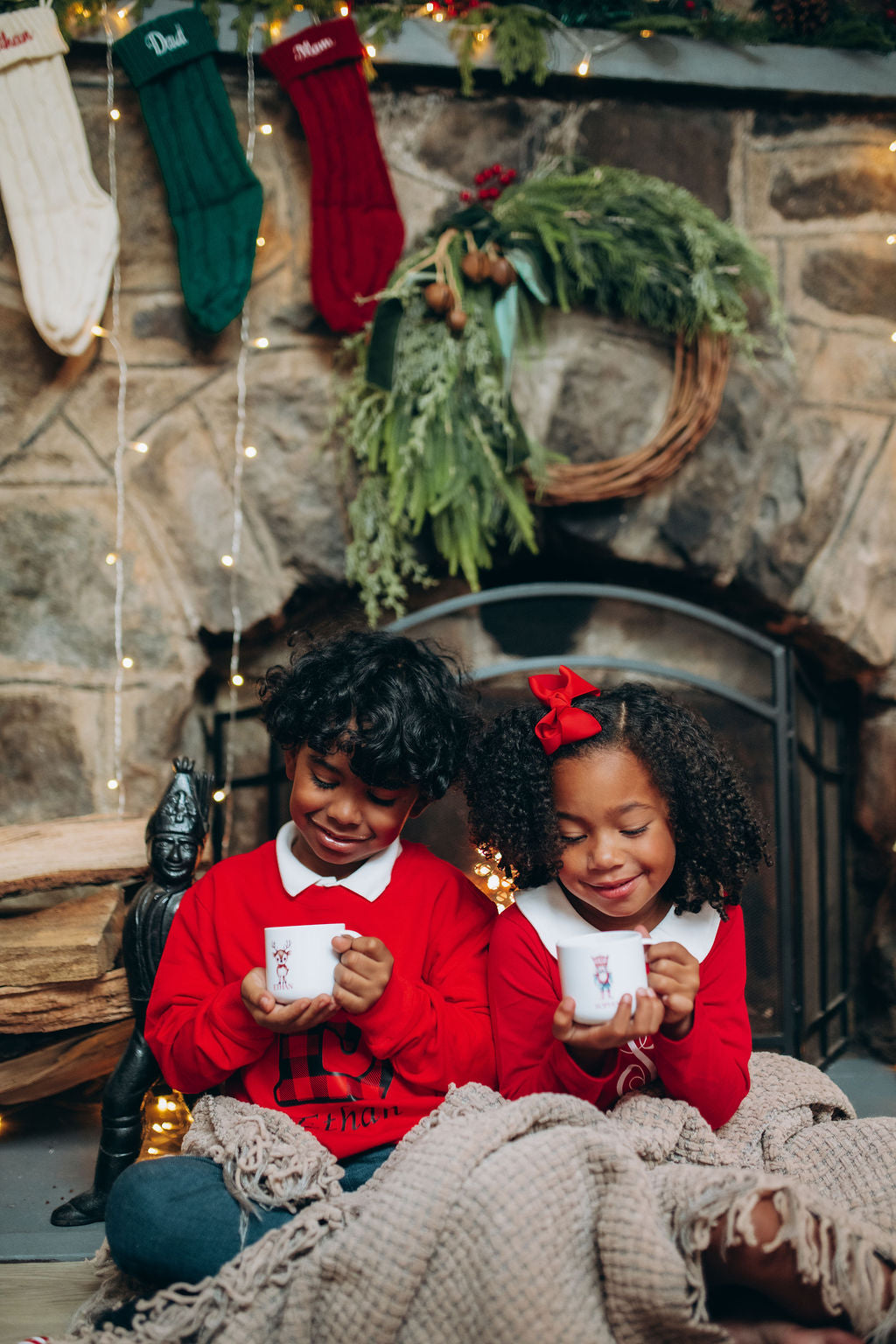 Two children sitting by a fireplace holding small white cups, with Christmas stockings and decorations in the background.