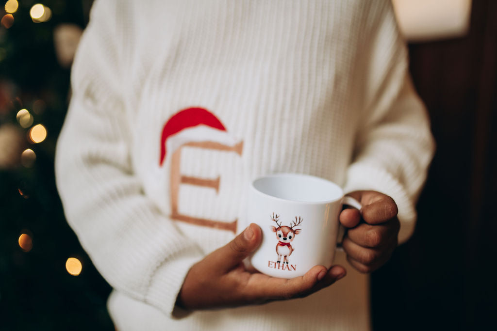 toddler wearing a festive sweater holding a mug with a reindeer design, Christmas tree in the background