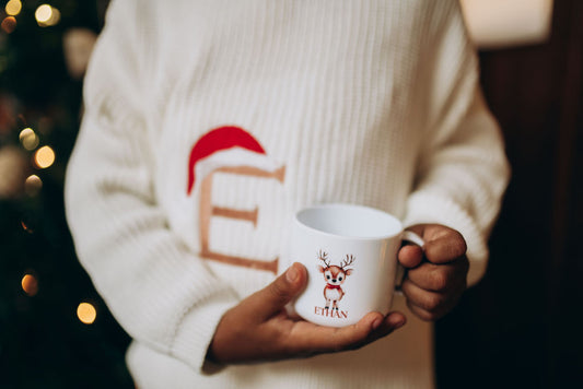 toddler wearing a festive sweater holding a mug with a reindeer design, Christmas tree in the background