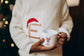 toddler wearing a festive sweater holding a mug with a reindeer design, Christmas tree in the background