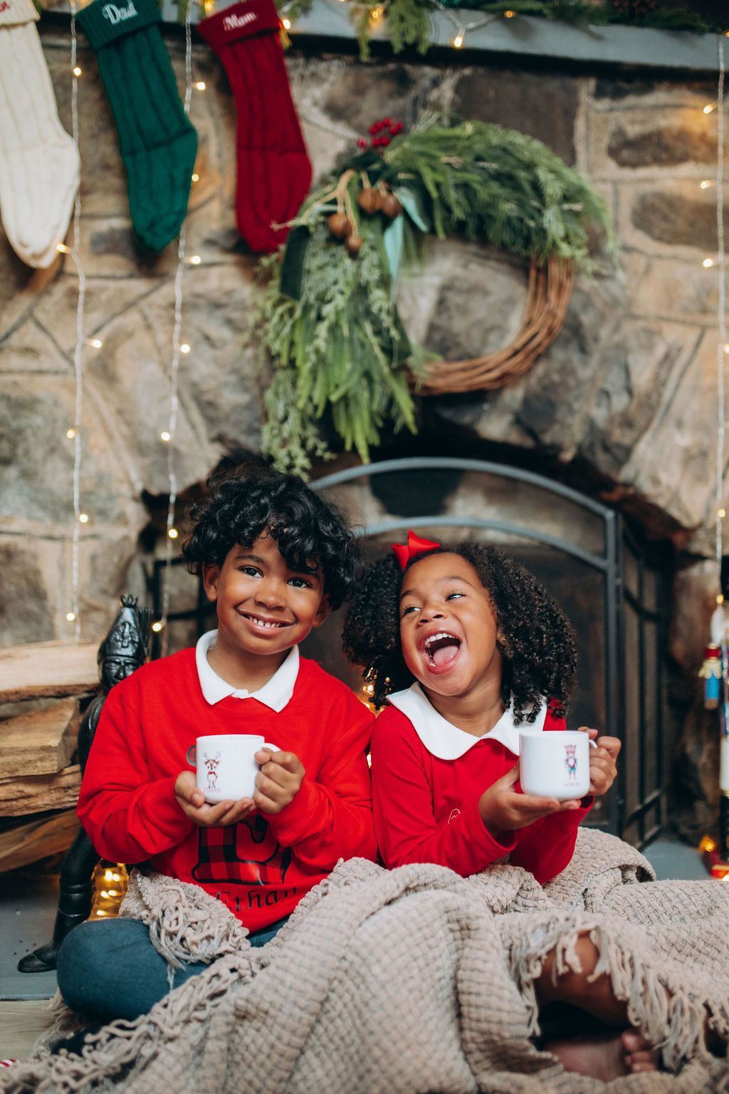 Two children in red sweaters holding mugs in front of a stone fireplace with Christmas decorations.