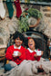 Two children in red sweaters holding mugs in front of a stone fireplace with Christmas decorations.