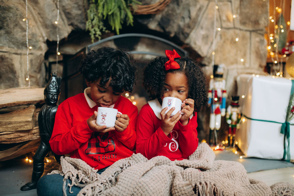 Two children in red sweaters holding mugs in front of a fireplace with Christmas decorations.