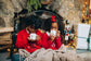 Two children in red sweaters holding mugs in front of a fireplace with Christmas decorations.